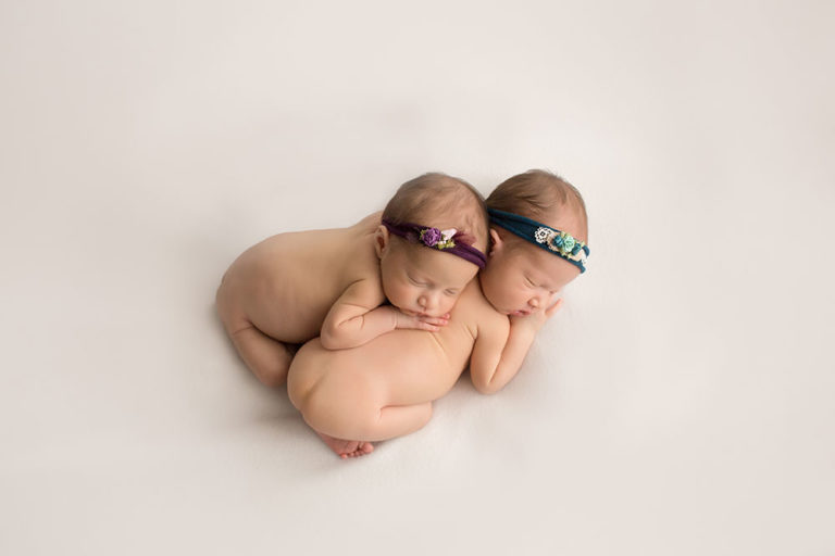 Newborn girls fraternal twins posed naked heads leaning against each other burgundy teal floral headbands posed on white blanket Gainesville FL baby photos