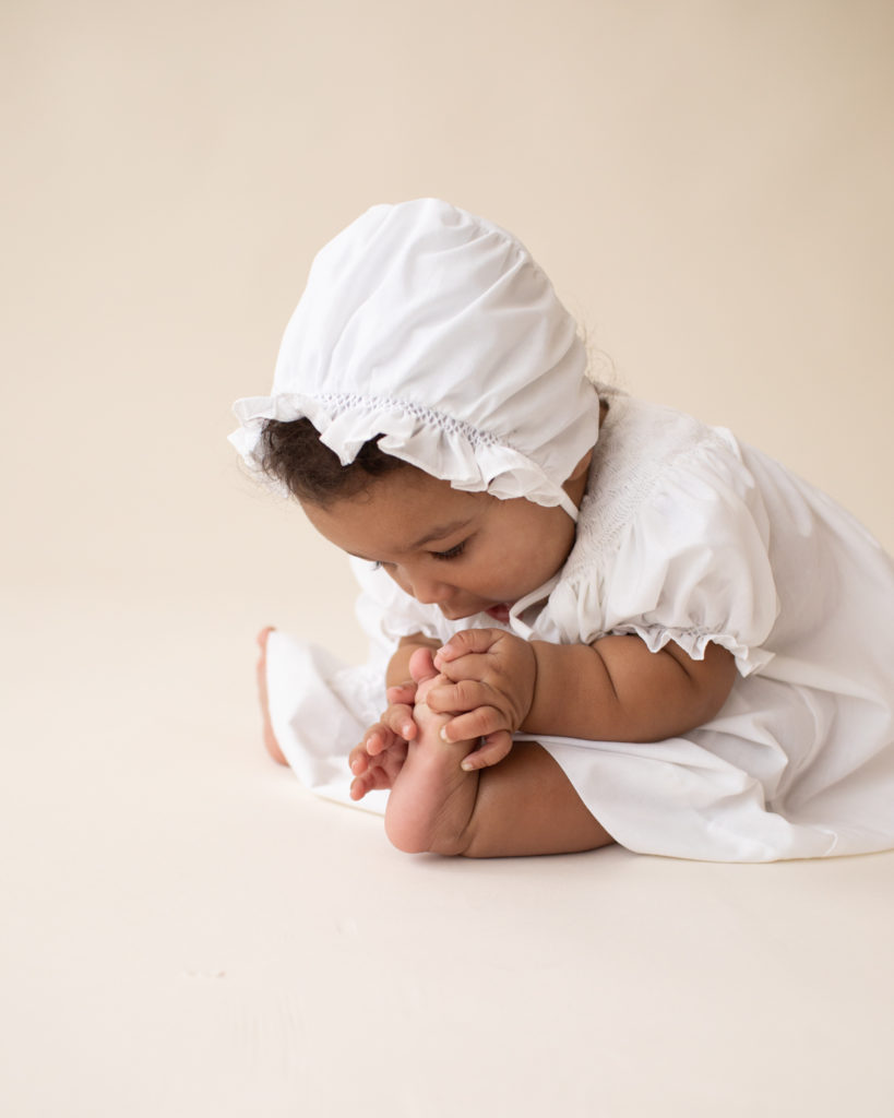 Baby 6 months old sitting up reaching for toes dressed in white cotton dress and matching bonnet Gainesville FL