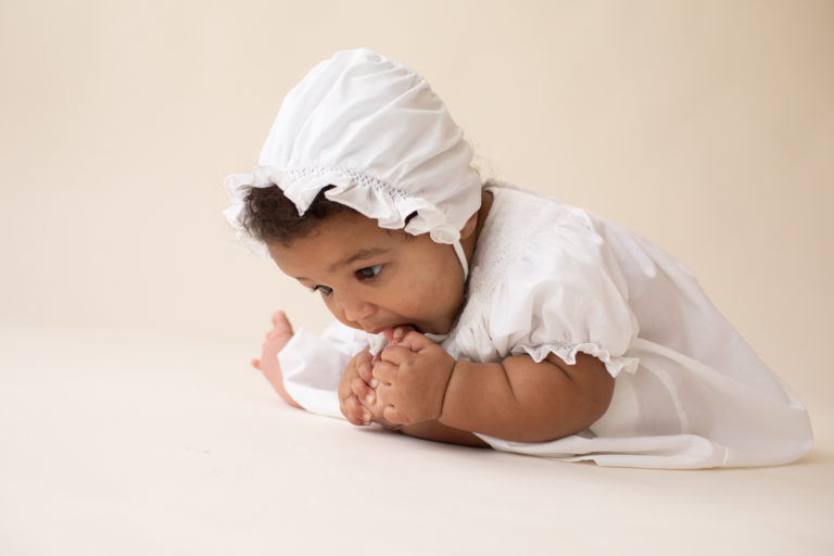 Baby 6 months old sitting up sucking her toes dressed in white cotton dress and matching bonnet Gainesville FL