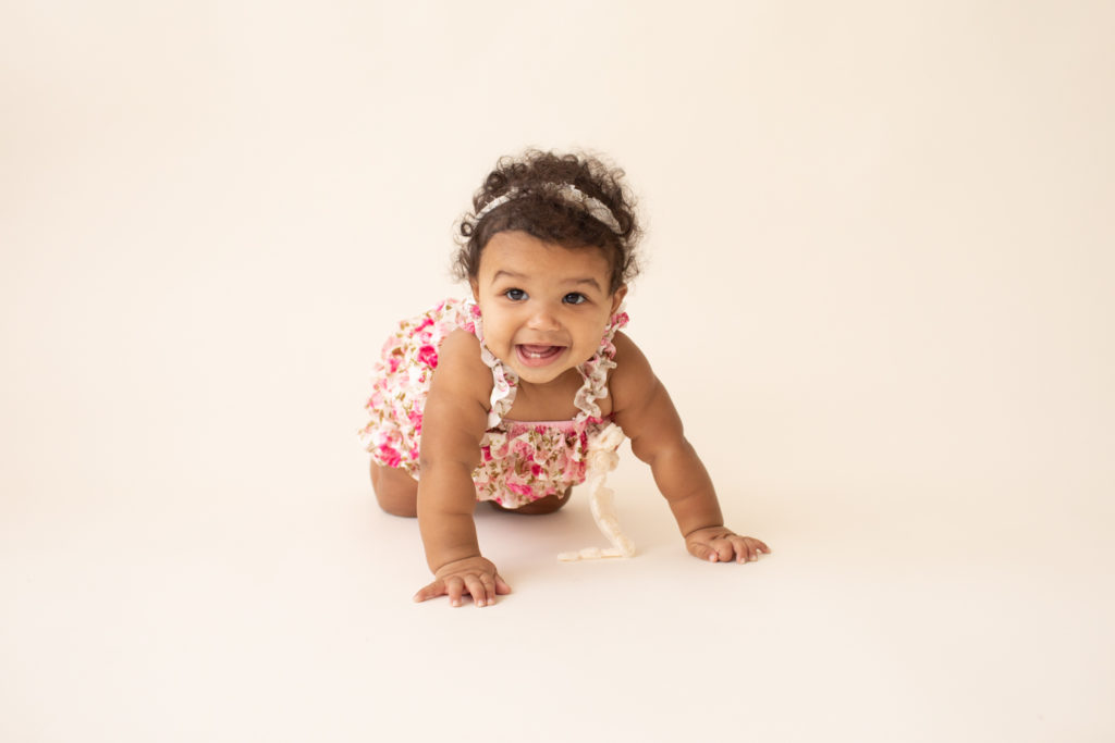 Baby 6 months old smiling crawling on white floor dressed in pink floral jumper in Gainesville FL