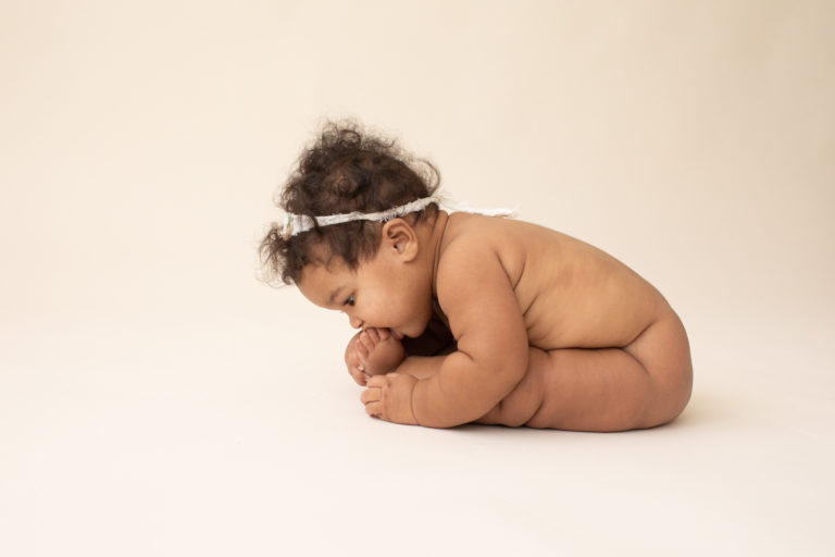 Milestone session 6 months old smiling naked with soft squishy baby rolls sucking toes on white floor in Gainesville FL