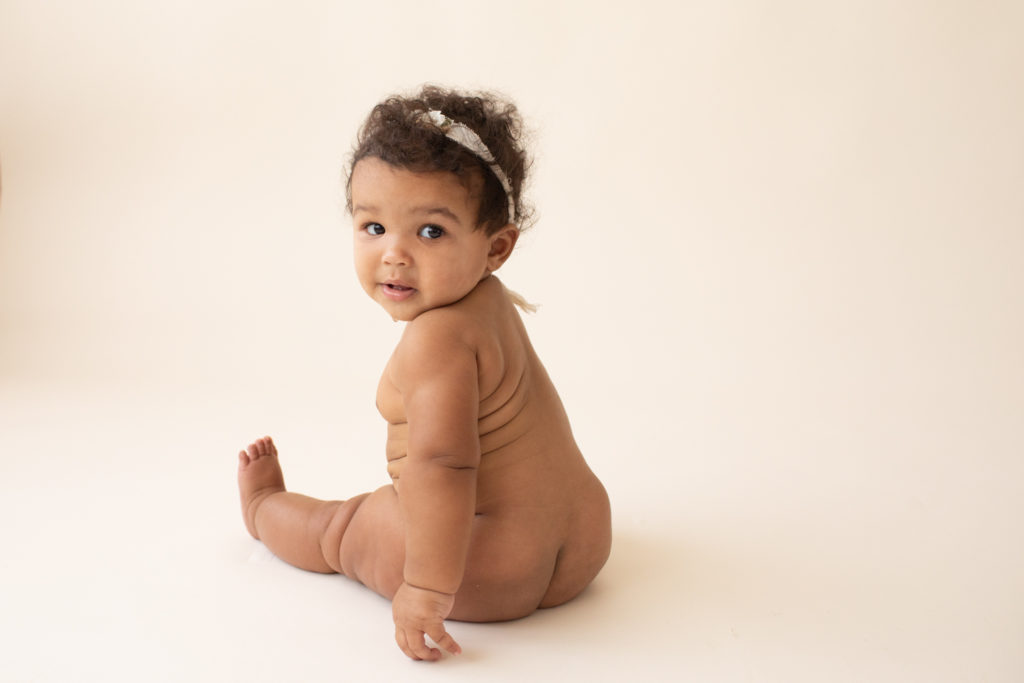 Milestone session 6 months old smiling sitting naked with soft squishy baby rolls glancing over shoulder to camera on white floor in Gainesville FL