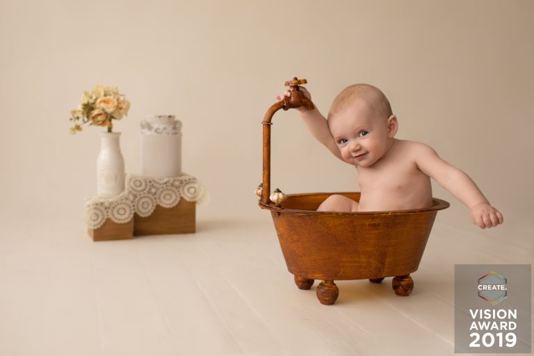 Award winning newborn photography baby posed in brown vintage tub with a teasing smile at floral and lace accents against ivory background