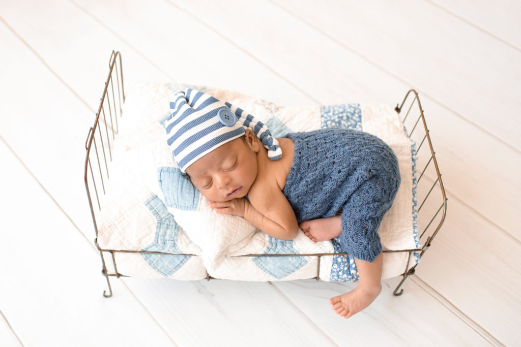 Newborn Jacob photos in blue knit overalls with a striped sleepy hat posed on quilt covered metal bed Gainesville FL