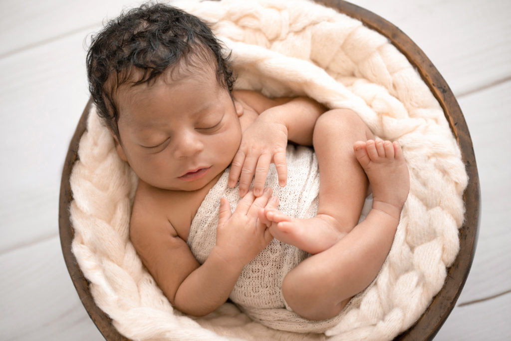 Newborn Jacob photos with full head of hair wrapped in cream knit wrap on cream fur photographed from above in brown wood bowl against cream wood floor Gainesville FL