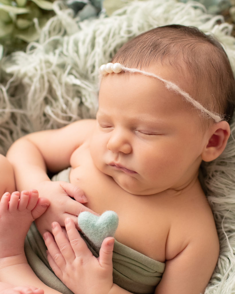 Hensley newborn photos close up tiny baby details sleeping in round fur textures basket surrounded with shades of green flowers baby hands holding tiny felted green heart from above Gainesville Florida Newborn Photography