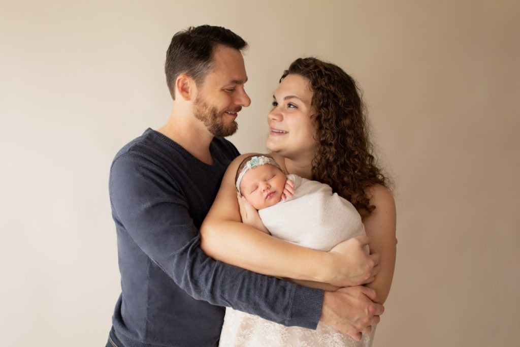 Hensley newborn photos proud Dad and delighted Mom dressed in ivory lace holding tiny swaddled baby in beige wrap smiling at each other Gainesville Florida Newborn Photography