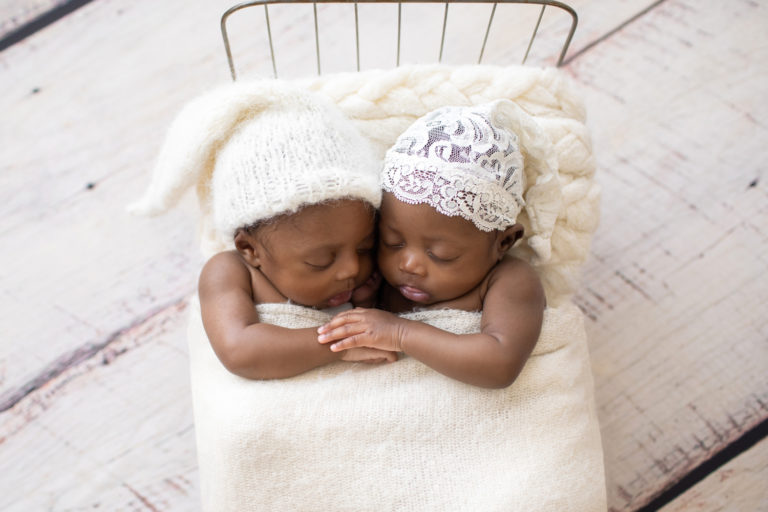 Twin Newborns Samuel and Sophia posed cuddling holding hands in metal newborn bed covered with cream blanket sleepy hats textured pillow distressed white floor in Gainesville Florida newborn photos taken from above