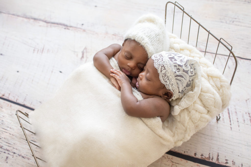 Twin Newborns Samuel and Sophia posed cuddling holding hands in metal newborn bed covered with cream blanket sleepy hats textured pillow distressed white floor in Gainesville Florida backlight newborn photos