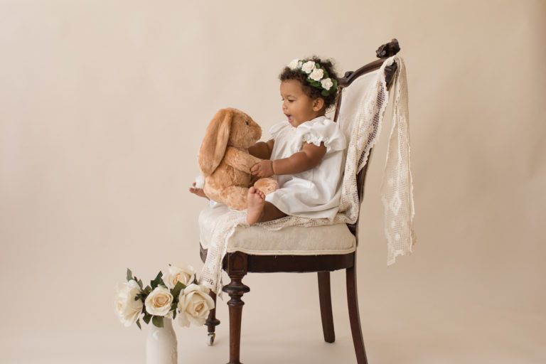 One year old Sara sitting with stuffed bunny on ivory victorian chair with lace smocked white dress milestone session Gainesville Florida