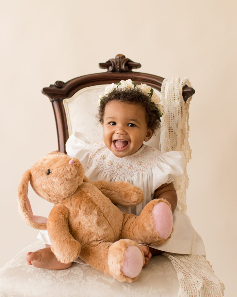 One year old Sara close up smiling sitting with stuffed bunny on ivory victorian chair with lace smocked white dress milestone session Gainesville Florida
