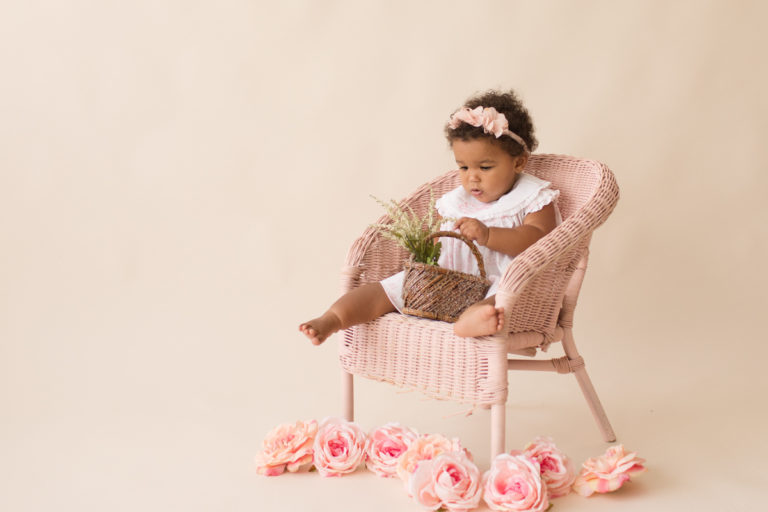 One year old Sara sitting pink wicker chair flower basket smocked white dress floral crown pink flowers on floor Gainesville Florida