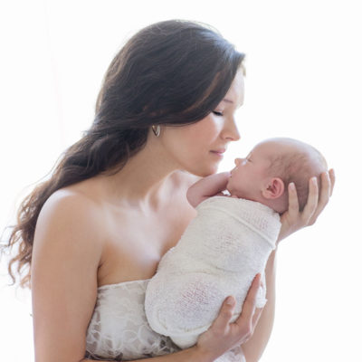 Young mom in lace adoring her newborn girl wrapped in white with backlight