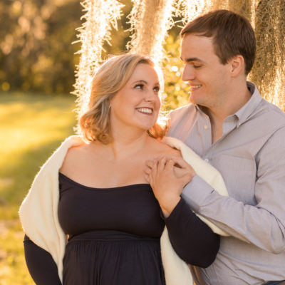 Maternity couple outdoors framed by moss draped sun lit tree Katie in navy blue maternity gown with white shawl and husband Ben in denim and grey Gainesville Florida Maternity portraits