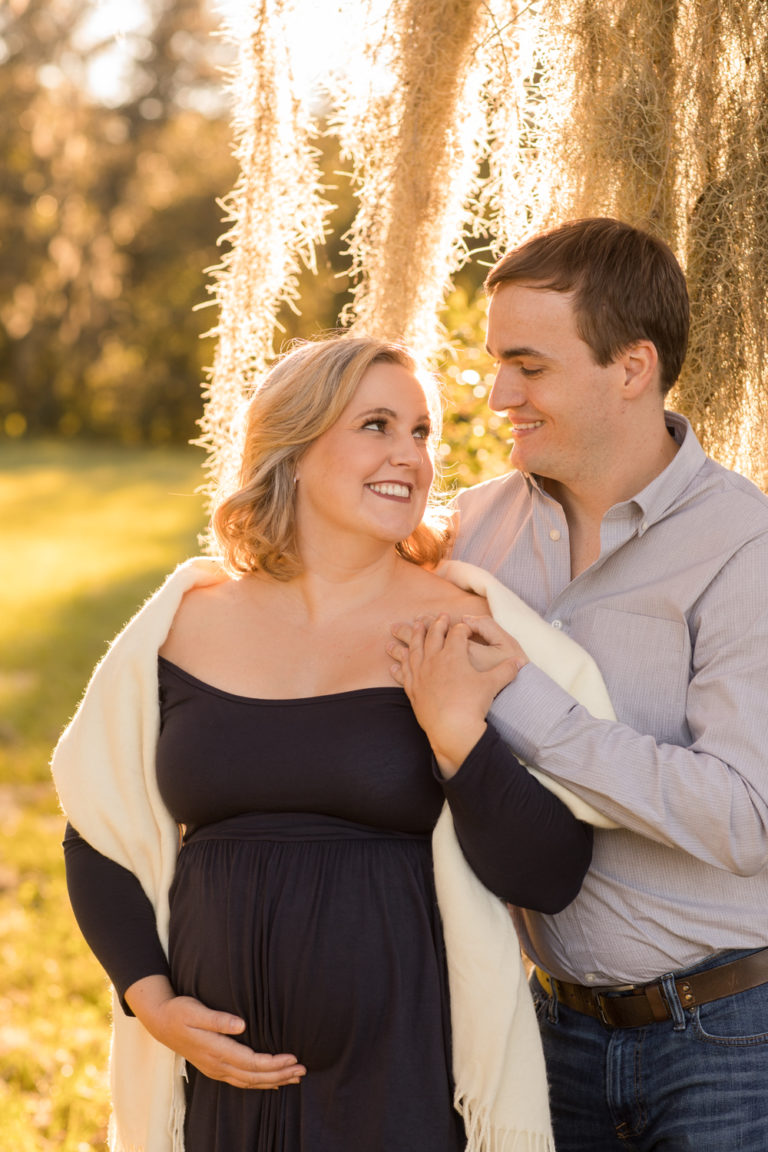 Maternity couple outdoors framed by moss draped sun lit tree Katie in navy blue maternity gown with white shawl and husband Ben in denim and grey Gainesville Florida Maternity portraits