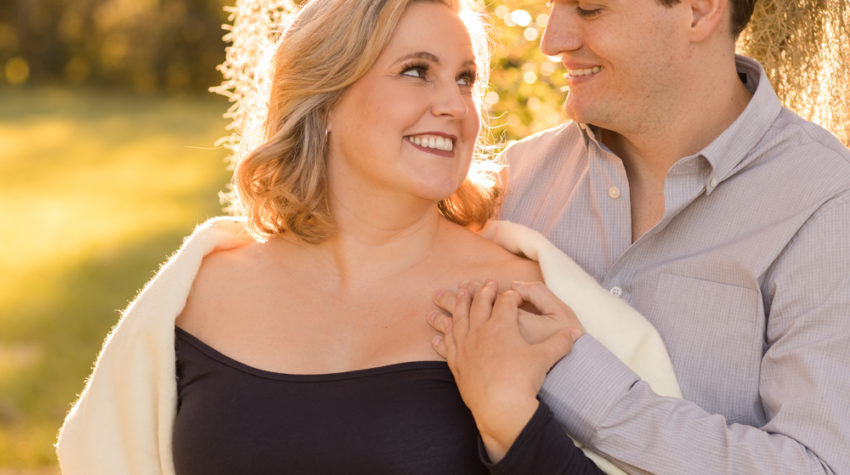 Maternity couple outdoors framed by moss draped sun lit tree Katie in navy blue maternity gown with white shawl and husband Ben in denim and grey Gainesville Florida Maternity portraits