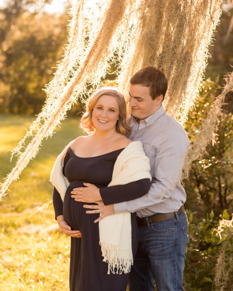 Maternity couple framed by outdoors moss draped sun lit tree Katie in navy blue maternity gown with white shawl and husband Ben in denim and grey admiring pregnant wife holding perfect baby bump Gainesville Florida Maternity portraits