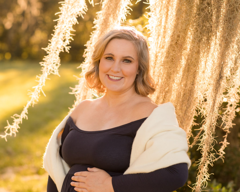 Maternity beauty Katie outdoors framed by moss draped sun lit tree in navy blue maternity gown with white shawl holding perfect baby bump Gainesville Florida Maternity portraits