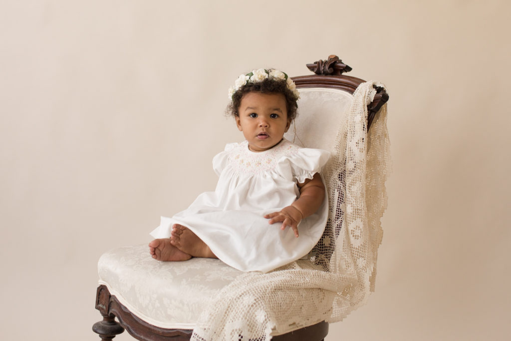 One year baby pictures Sara wearing white heirloom smocked dress and white floral crown posed sitting on lace draped elegant ivory chair with cream background Gainesville Florida Baby Photography