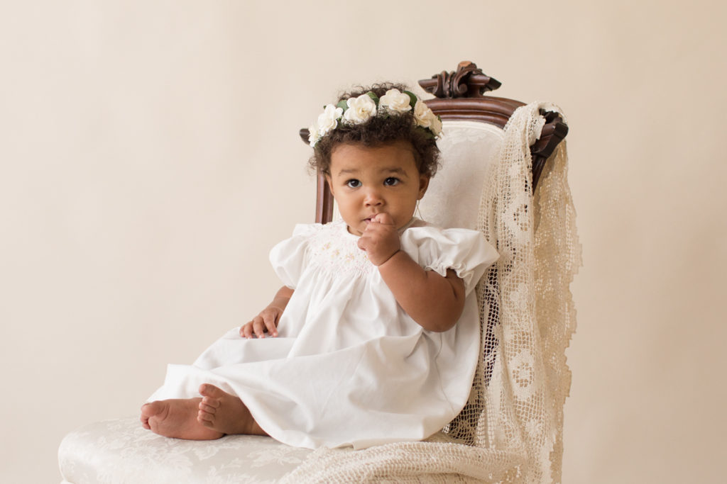 One year baby pictures Sara wearing white heirloom smocked dress and white floral crown shy look posed sitting on lace draped elegant ivory chair with cream background Gainesville Florida Baby Photography