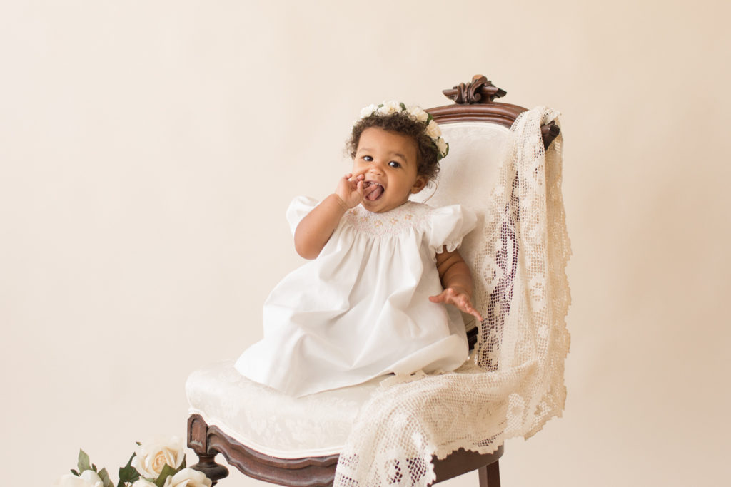 One year baby pictures Sara wearing white heirloom smocked dress and white floral crown sucking fingers smiling posed sitting on lace draped elegant ivory chair with cream background Gainesville Florida Baby Photography