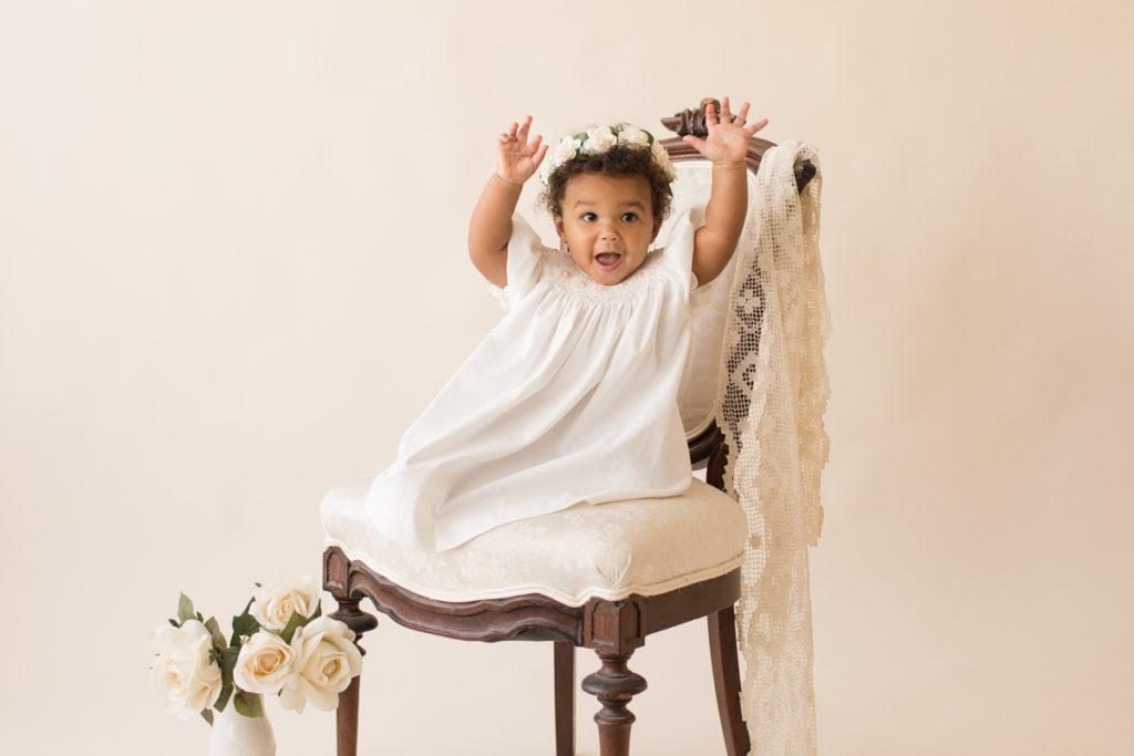 One year baby pictures Sara wearing white heirloom smocked dress and white floral crown excited happy arms straight up in air posed sitting on lace draped elegant ivory chair with white roses cream background Gainesville Florida Baby Photography