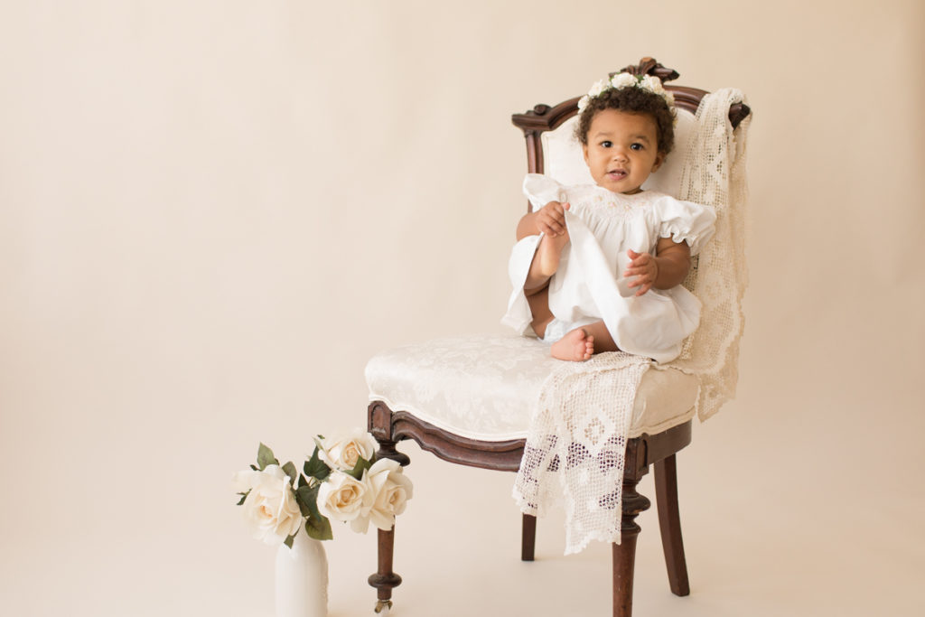 One year baby pictures Sara wearing white heirloom smocked dress and white floral crown excited holding foot posed sitting on lace draped elegant ivory chair with white roses cream background Gainesville Florida Baby Photography