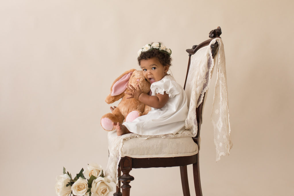 One year baby pictures Sara wearing white heirloom smocked dress and white floral crown excited hugging stuffed bunny posed profile sitting on lace draped elegant ivory chair with white roses cream background Gainesville Florida Baby Photography