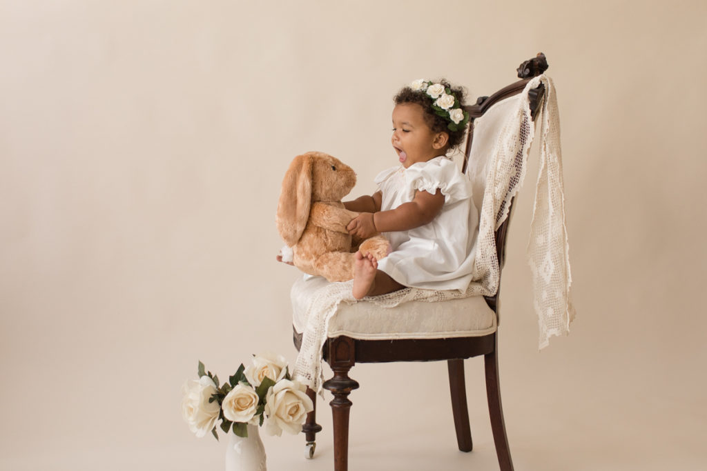 One year baby pictures Sara wearing white heirloom smocked dress and white floral crown excited talking to stuffed bunny posed sitting profile on lace draped elegant ivory chair with white roses cream background Gainesville Florida Baby Photography