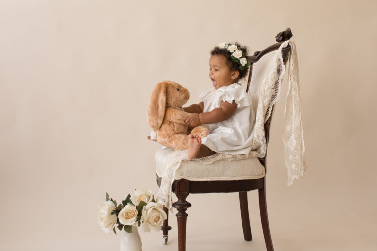 One year baby pictures Sara wearing white heirloom smocked dress and white floral crown excited talking to stuffed bunny posed sitting profile on lace draped elegant ivory chair with white roses cream background Gainesville Florida Baby Photography