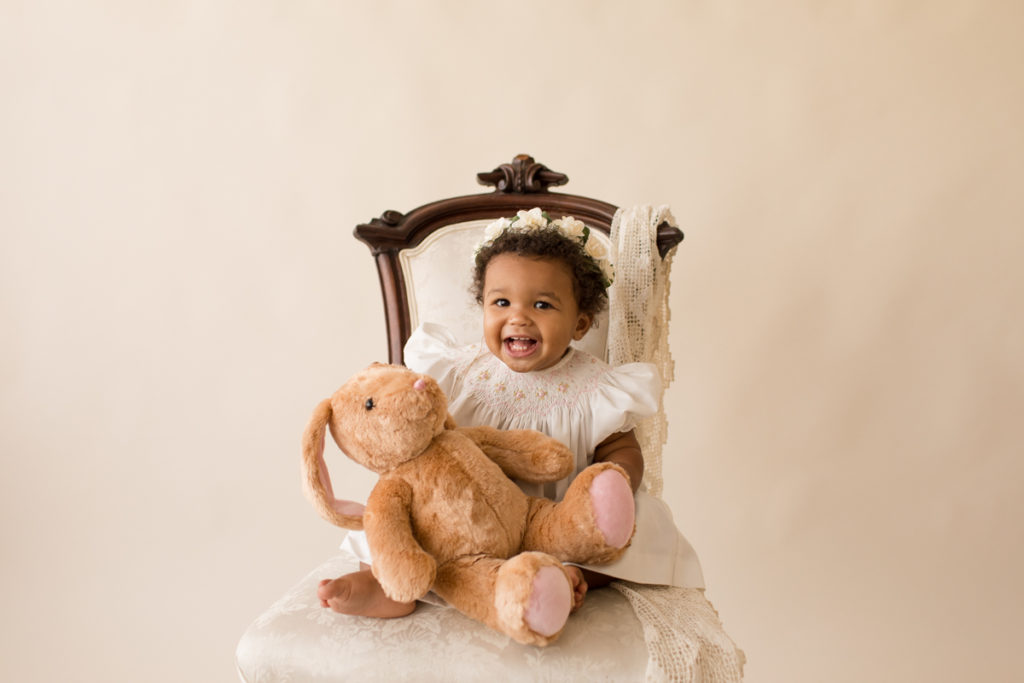 One year baby pictures Sara wearing white heirloom smocked dress and white floral crown excited holding stuffed bunny big smiles posed sitting on lace draped elegant ivory chair with cream background Gainesville Florida Baby Photography