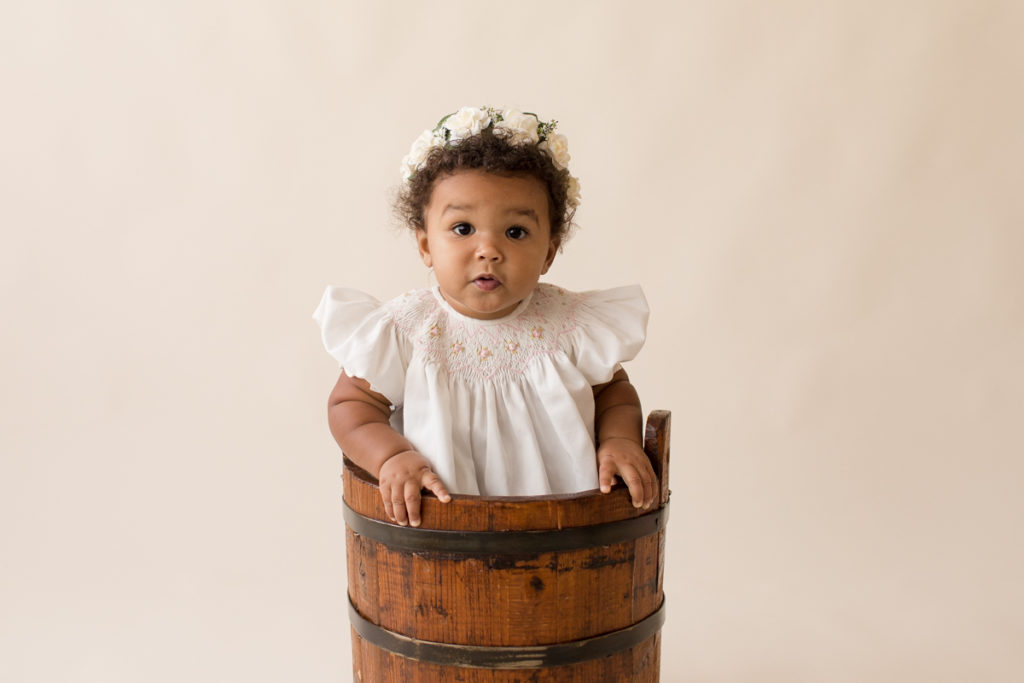 One year baby pictures Sara wearing white heirloom smocked dress and white floral crown sitting in wooden bucket cream background Gainesville Florida Baby Photography