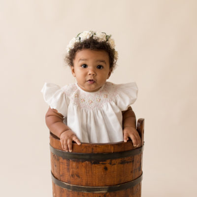 One year baby pictures Sara big brown eyes and baby rolls wearing white heirloom smocked dress and white floral crown sitting in wooden bucket cream background Gainesville Florida Baby Photography