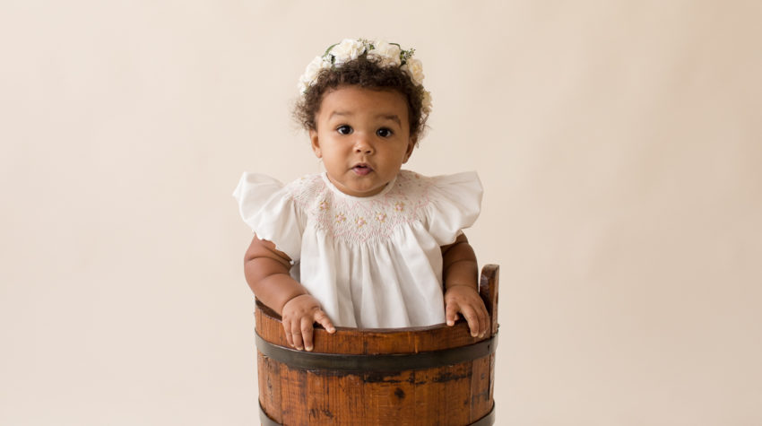 One year baby pictures Sara big brown eyes and baby rolls wearing white heirloom smocked dress and white floral crown sitting in wooden bucket cream background Gainesville Florida Baby Photography