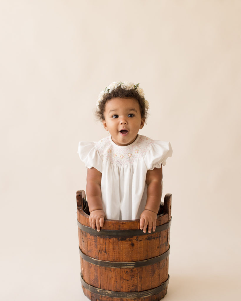 One year baby pictures Sara wearing white heirloom smocked dress and white floral crown standing up in wooden bucket cream background Gainesville Florida Baby Photography