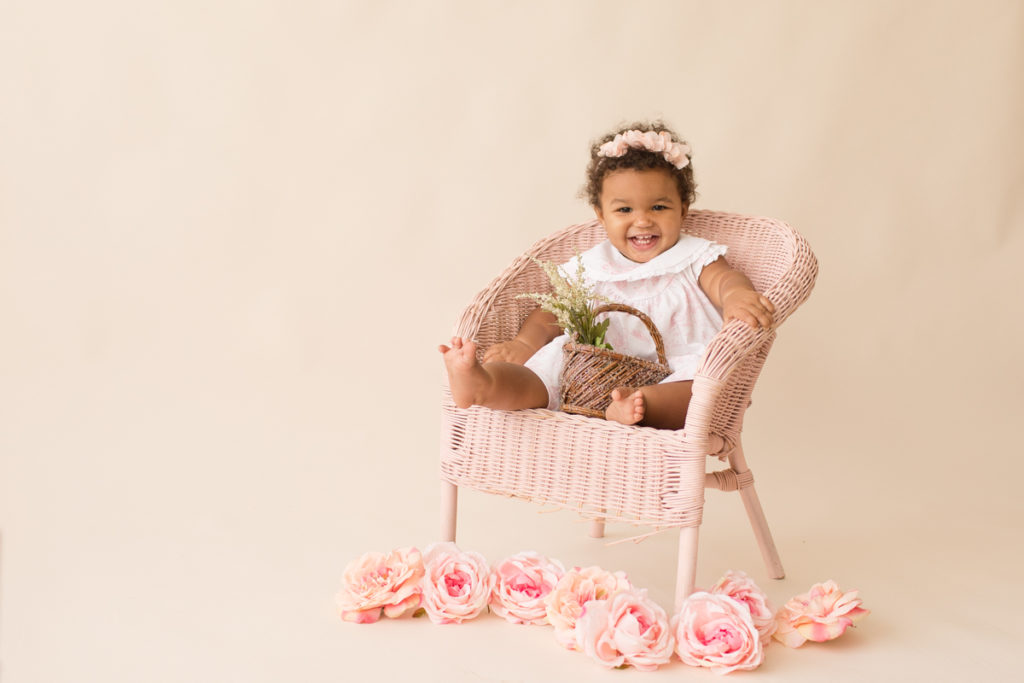 One year baby pictures Sara wearing white heirloom smocked dress and pink floral crown big smile holding tiny basket of flowers posed sitting on pink wicker children chair with pink and peach roses cream background Gainesville Florida Baby Photography