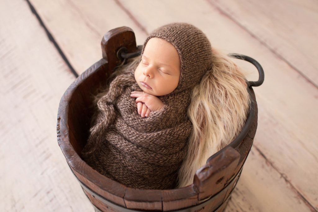 Adorable Nathan sleeping in fur stuffed brown wooden bucket all wrapped up in a brown knit potato sack with brown bonnet photo from side view newborn portrait