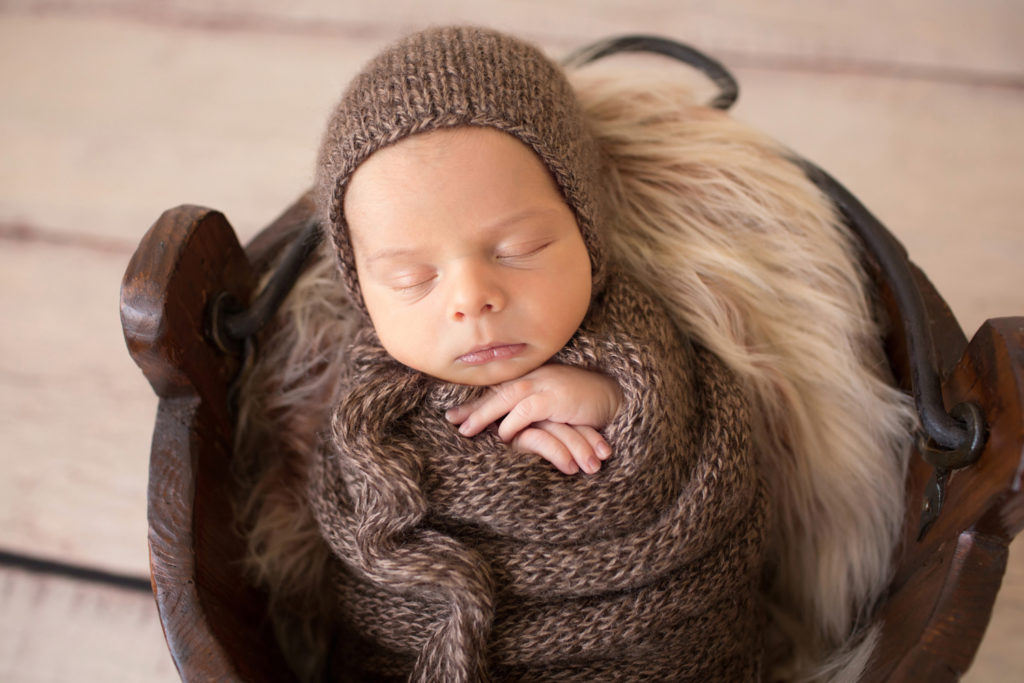 Adorable newborn baby sleeping in fur stuffed brown wooden bucket all wrapped up in a brown knit potato sack with brown bonnet newborn portrait