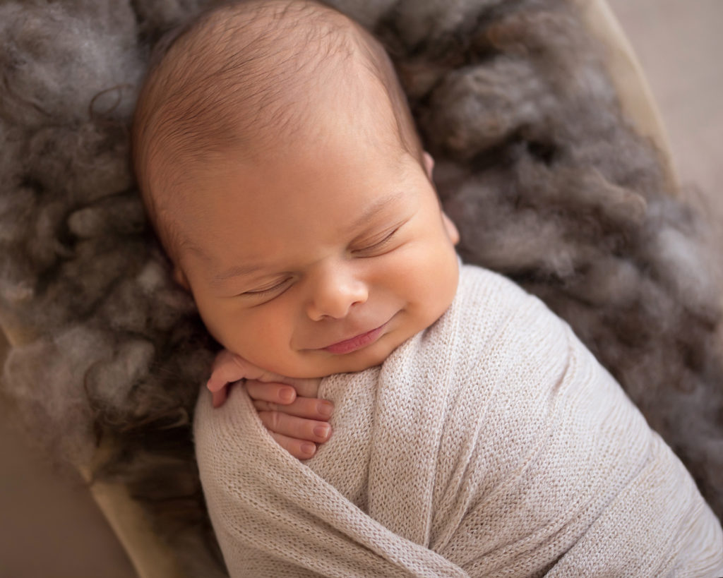 Newborn baby boy Nathan big baby smile in cream newborn wrap posed on brown wool in a cradle close up newborn portrait