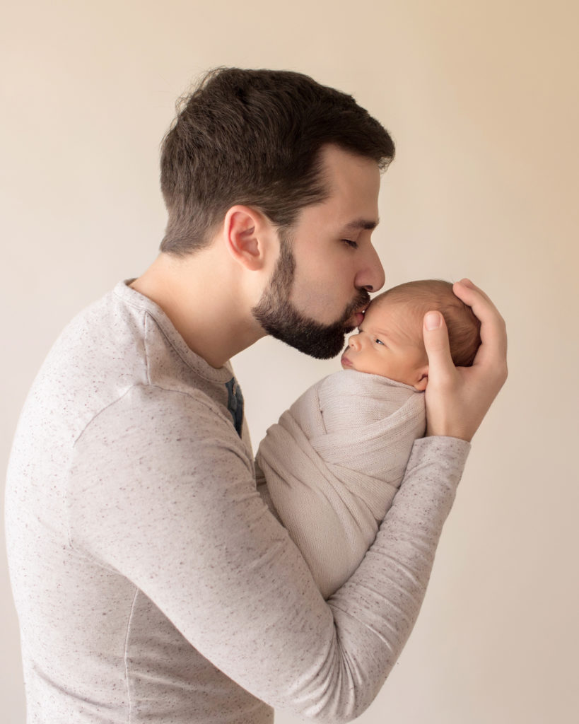 Handsome dad tenderly kissing newborn boy wrapped in cream soft neutral colors profile photo baby with open eyes Gainesville FL newborn photography