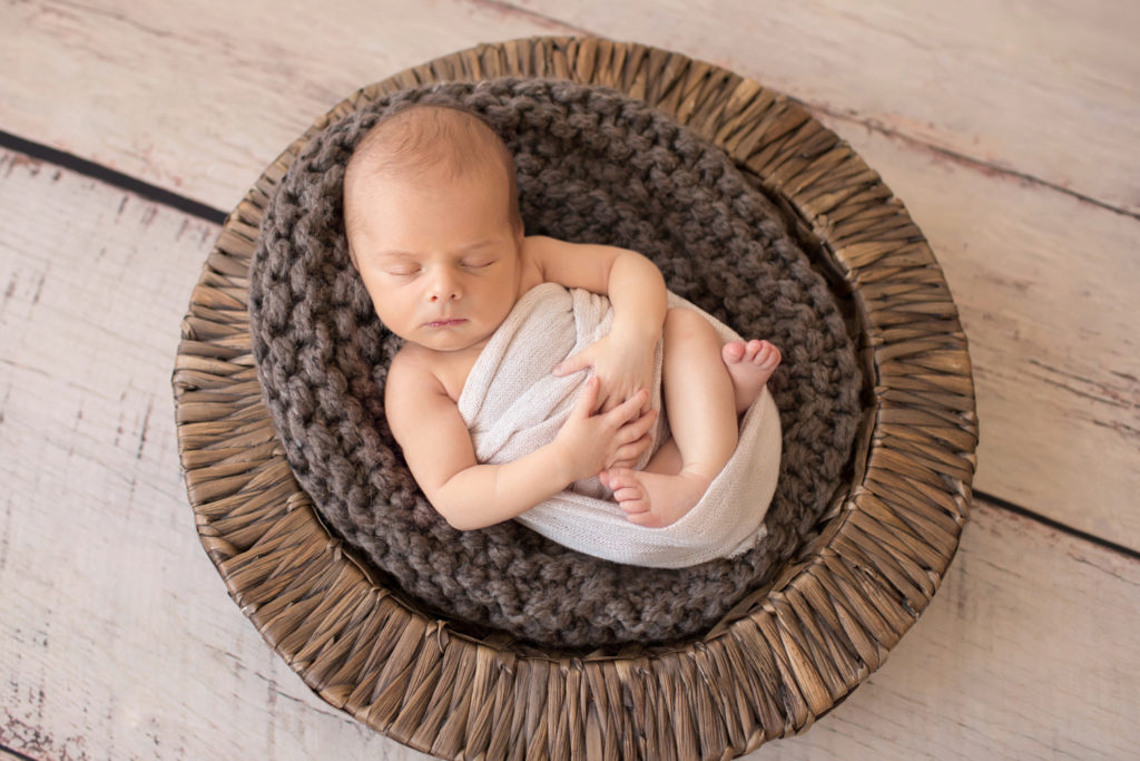 Newborn boy Nathan swaddled in cream wrap posed in round brown basket stuffed with chunky knit brown blanket newborn portrait