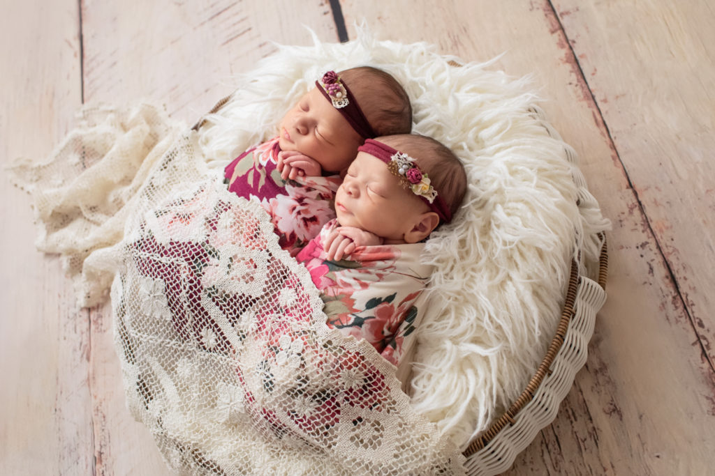 Newborn twin girls photos Renae and Janae in rose and burgundy floral wraps and floral head ties in a fur stuffed round basket draped in lace angle shot