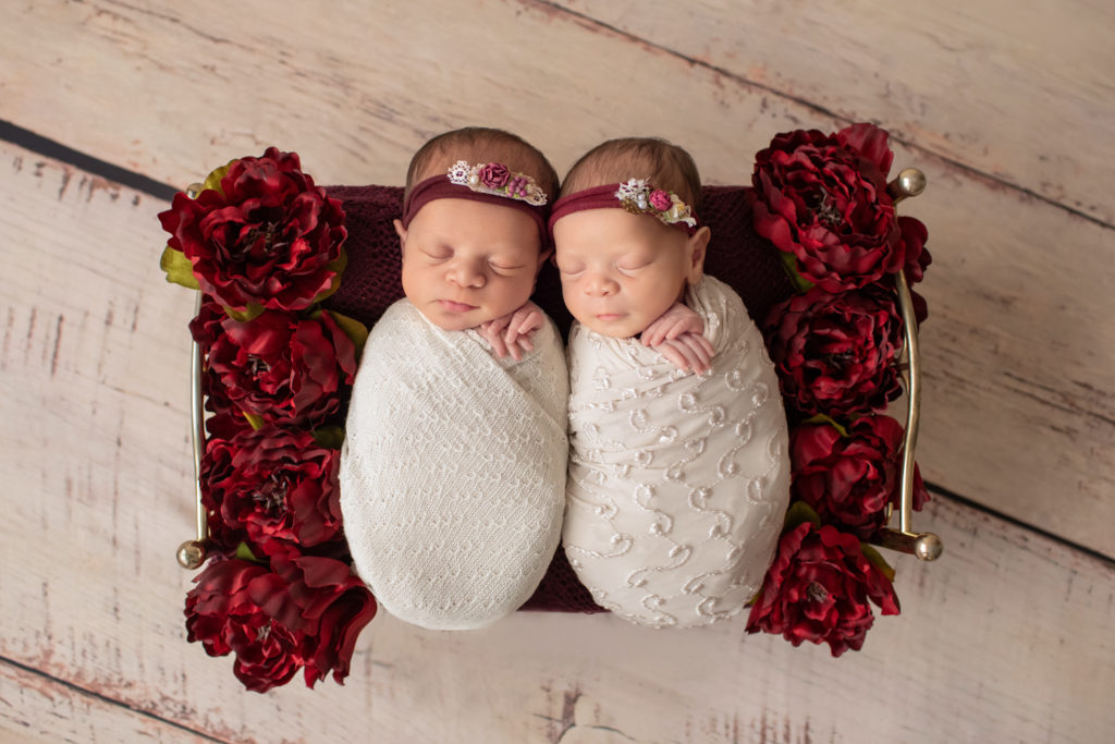 Newborn twin girls photos swaddled in delicate cream wrap and floral head tie lying on a Christmas brass bed surrounded by burgundy peonies to their left and right on white washed wood floor
