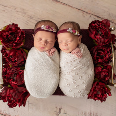 Newborn twin girls photos swaddled in delicate cream wrap and floral head tie lying on a Christmas brass bed surrounded by burgundy peonies to their left and right on white washed wood floor