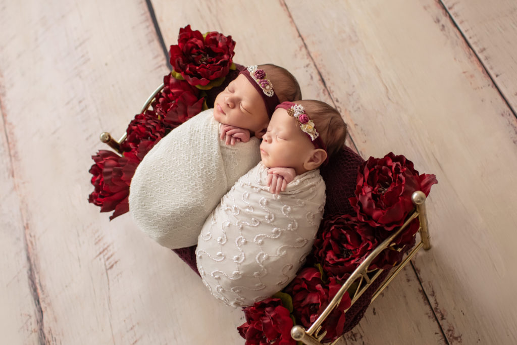Newborn twin girl photos swaddled in delicate cream wrap and floral head ties lying on a Christmas brass bed surrounded by burgundy peonies to their left and right angled shot