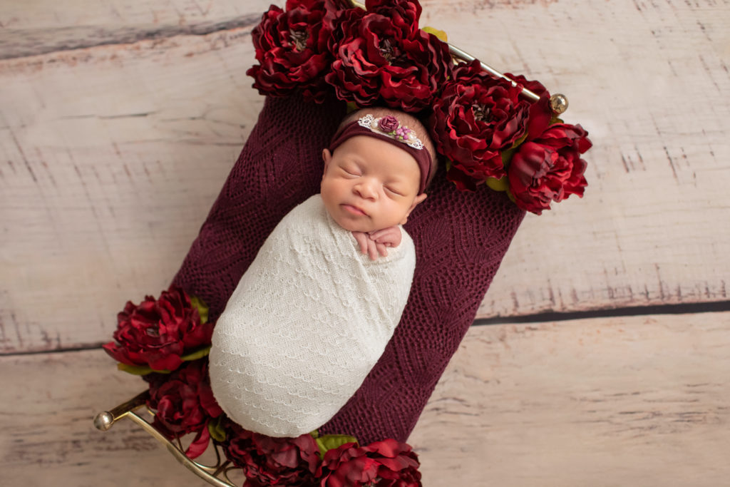 Newborn girl photo swaddled in delicate cream wrap and floral head tie lying on a Christmas brass bed surrounded by burgundy peonies at top and bottom