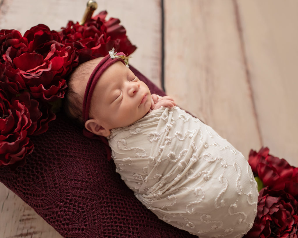 Newborn girl photo swaddled in delicate cream wrap and floral head tie lying on a Christmas brass bed surrounded by burgundy peonies to their left and right