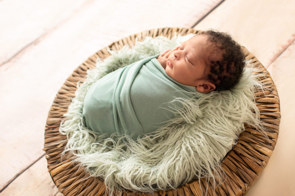 Newborn boy 2 weeks old lots of curly hair sage green wrap in fur stuffed basket backlit