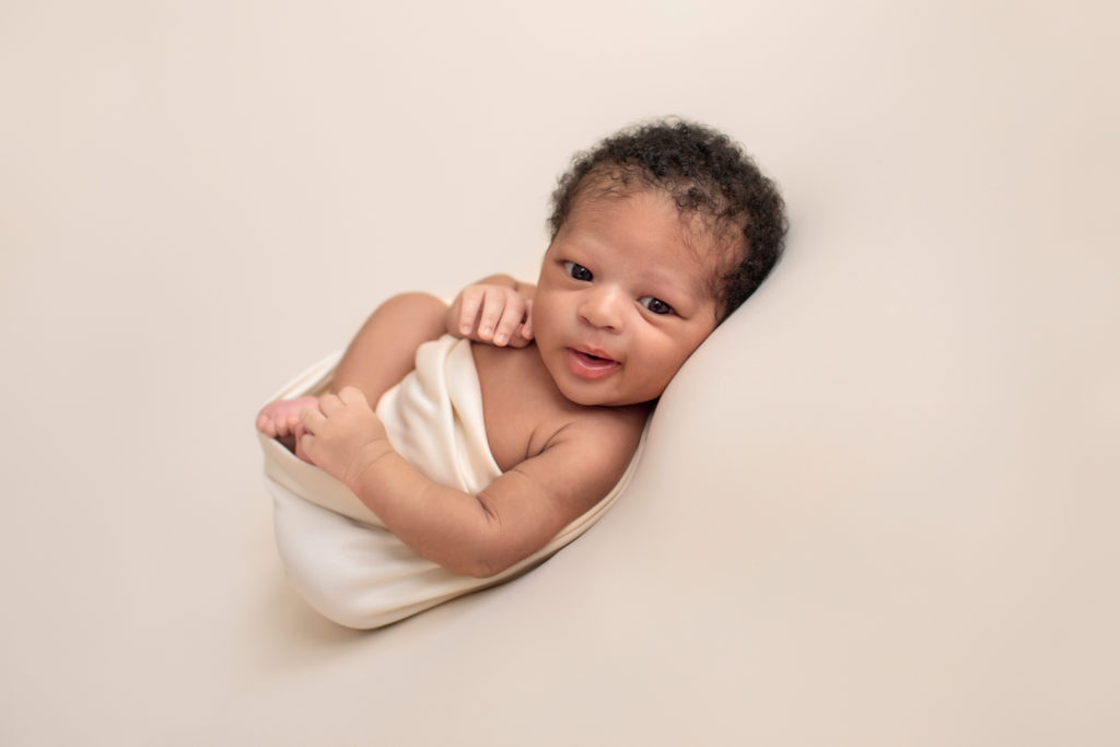 Gainesville newborn photos baby posed on his back on neutral cream blanket big brown eyes wide open baby Chad grinning at camera head full of curly brown hair