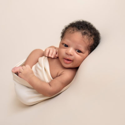 Gainesville newborn photos baby posed on his back on neutral cream blanket big brown baby eyes wide open baby Chad grinning at camera head full of curly brown bsby hair